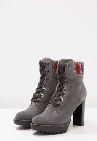 Bottes chelsea en daim gris avec lacets texturés foncés, un revers en tissu à motifs rouge et gris, et un talon épais noir. Semelle en caoutchouc à crampons.