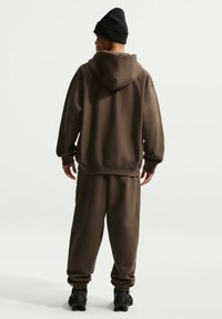 Hombre vestido con sudadera con capucha marrón oscuro, pantalones deportivos a juego, gorro negro y zapatillas negras, de pie de espaldas a la cámara sobre un fondo blanco liso.