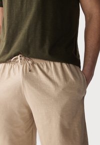 Beige elastic pants with drawstring waist, paired with a dark green short-sleeve shirt. Soft, textured fabric in relaxed fit.