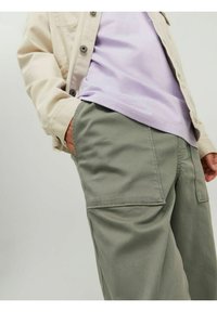 Giacca button-up beige chiaro sopra una maglietta lavanda e pantaloni cargo verdi, con tasche laterali e una vestibilità rilassata.