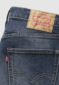 Τζιν παντελόνι με σκουρό μπλε χρώμα, κίτρινη ραφή και vintage ετικέτα Levi's με λεπτομέρειες προϊόντος. Η πίσω τ σέπη εμφανίζει το εμβληματικό λογότυπο.