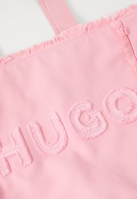 Borsa a tracolla in tela rosa con bordi sfilacciati, caratterizzata da un ricamo testurizzato della parola "HUGO" in filo rosa chiaro.