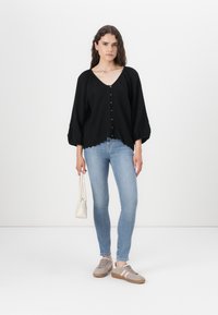 Black, textured blouse with a V-neck and front buttons, paired with light blue skinny jeans and beige sneakers. Also holding a small white bag.