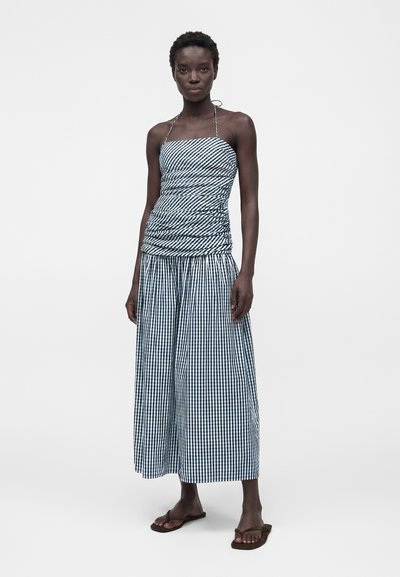 Γυναίκα με μαύρο-άσπρο καρό φόρεμα με χαλαρή μπούστα και φαρδιά φούστα, όρθια ξυπόλυτη πάνω σε λευκό φόντο.