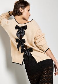 Suéter beige hecho de material suave. Presenta tres lazos de satén negro en la parte posterior y un borde negro contrastante. Se lleva sobre un tejido de encaje negro.
