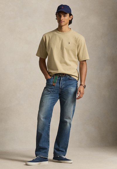 Jeune homme portant un t-shirt beige, un jean bleu avec une ceinture rayée, une casquette marine et des baskets bleues, debout avec une main dans la poche.