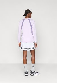 Ανοιχτό μωβ μπλούζα με μακριά μανίκια και αντίθετο πίσω πάνελ σε σκούρο μωβ, συνδυασμένη με λευκά σορτς με μαύρες ρίγες και γκρι κάλτσες.