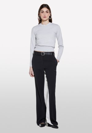 Donna che indossa una maglia grigio chiaro a maniche lunghe, pantaloni neri a gamba larga con una striscia bianca, cintura nera, scarpe nere e grandi orecchini a cerchio.
