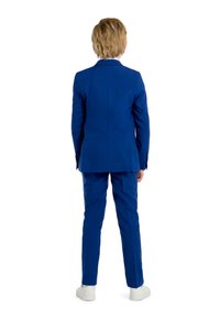 Giacca e pantaloni blu realizzati in un tessuto liscio, caratterizzati da una vestibilità su misura. Sneakers bianche completano l'outfit. Vista da dietro.