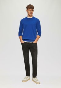 Pull bleu à col rond avec des manches longues, porté sur une chemise blanche, associé à un jean noir et des baskets gris clair avec des semelles beiges.