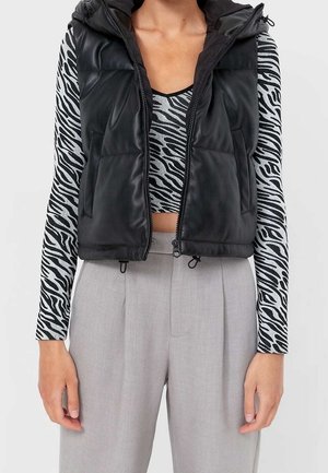 Personne portant un gilet matelassé noir sans manches sur un haut à manches longues imprimé zèbre, associé à un pantalon taille haute gris clair.