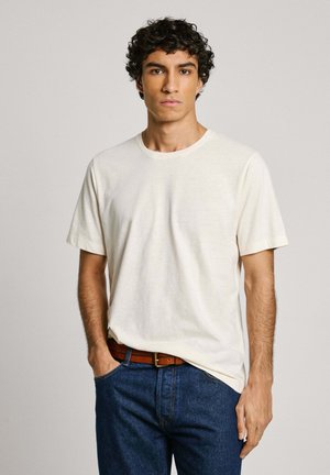 Camiseta de algodón color crema con cuello redondo y mangas cortas, combinada con jeans de mezclilla azules y un cinturón de cuero marrón.