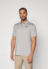 Gaiši pelēks Nike polo krekls ar īsām piedurknēm, apkakli, trim pogām un melnu logotipu kreisajā krūtī. Gluda tekstūra.