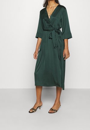 Mujer con un vestido cruzado de satén verde oscuro con mangas tres cuartos, cintura atada, sandalias de tacón con estampado de leopardo y collares dorados en capas.