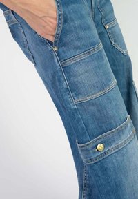 Pantalon cargo en denim bleu clair avec plusieurs poches, accents en métal doré et finition texturée. Présente des coutures visibles et un design de jambe fuselée.