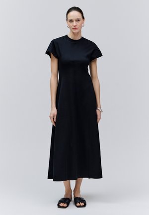 Frau steht in einem schwarzen ärmellosen Midikleid mit rundem Ausschnitt und schwarzen Slide-Sandalen vor einem schlichten hellgrauen Hintergrund.