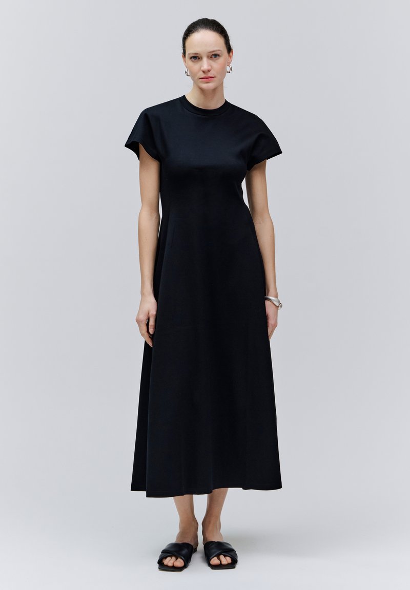 Femme debout dans une robe midi noire sans manches avec un col rond et des sandales noires à enfiler, sur un fond gris clair uni.