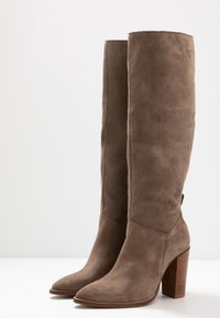 Botas marrones hasta la rodilla hechas de ante, con punta afilada y tacón apilado de madera, con una textura suave y detalles mínimos de costuras.