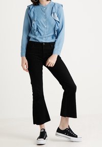 Camicia button-up azzurro chiaro con dettagli a rouches, abbinata a pantaloni a corduroy neri cropped a zampa di elefante e sneakers in tela nere.
