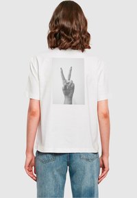 Weißes T-Shirt mit einem Grafikdruck einer Hand, die ein Peace-Zeichen macht, auf dem Rücken. Kurze Ärmel, lockere Passform und sichtbare Denim-Jeans.