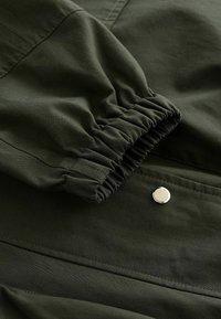 Tessuto verde scuro con polsino elastico arricciato e vistoso bottone a pressione argentato su un capo cucito.