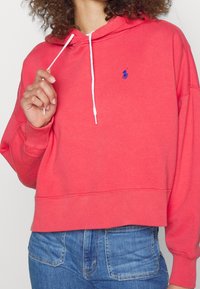 Korálová mikina s kapucí a stahovací šňůrkou, na prsou s malým modrým logem. Měkká látka, žebrované manžety a lem, volný střih, kombinována s modrými džíny.