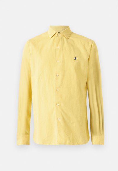 Κίτρινο πουκάμισο με μακρύ μανίκι φτιαγμένο από ύφασμα, διαθέτει κουμπιά μπροστά, αιχμηρό γιακά και ένα μικρό σκούρο λογότυπο στο αριστερό στήθος.