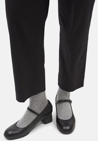 Black leather Mary Jane shoes with a rounded toe and block heel, paired with light gray ribbed socks, worn with black pants.