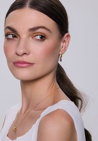 Złota kolczyk w kształcie obręczy z prostokątnym czarnym akcentem, o gładkim wykończeniu. Model nosi białą, teksturowaną bluzkę i delikatny złoty naszyjnik.