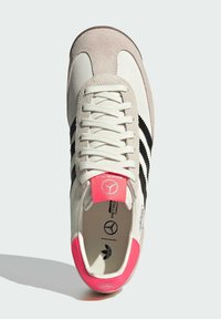 Bílé tenisky s černými pruhy, šedými surovými akcenty a růžovým patním poutkem. Mají tkaničky, logo na jazyku a strukturovaný povrch.