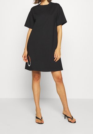 Mujer que lleva un vestido negro holgado de manga corta con un corte circular cerca del dobladillo, combinado con sandalias negras de tacón.
