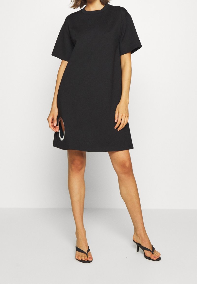 Femme portant une robe noire ample à manches courtes avec une découpe circulaire près de l'ourlet, associée à des sandales noires à talons.