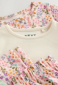 Zwei Oberteile flach ausgelegt. Eines ist aus cremefarbigem Rippstoff, das andere hat einen rosafarbenen Hintergrund mit einem mehrfarbigen Blumenmuster.