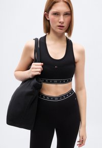 Μαύρο αθλητικό κορμάκι και κολάν με ζώνη μέσης με λογότυπο. Το μοντέλο κρατά μια μαύρη τσάντα ώμου που ταιριάζει, με απλό, εφαρμοστό σχέδιο.