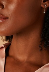 Des boucles d'oreilles en chaîne argentée présentent un accent de perle ronde, mettant en valeur un design délicat contre un cou lisse aux tons chauds et un haut en satin rose.