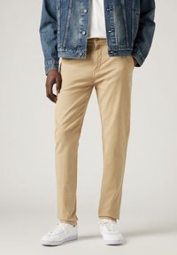 Chino beige con una vestibilità slim, caratterizzati da una texture liscia e tasche frontali, abbinati a una giacca di jeans blu e sneakers bianche.