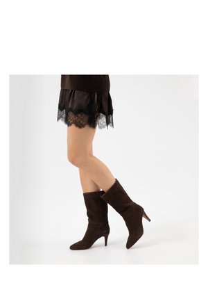 Donna che indossa una gonna nera con rifiniture in pizzo e stivali in suede marrone scuro con tacco alla metà del polpaccio, in piedi con le gambe incrociate su sfondo bianco.