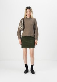 Brun strikket genser med bølget kant, kombinert med et grønt kordfløyelskjørt. Modellen holder en teksturert skulderveske med slangeskinnmønster. Svarte sko og sokker.