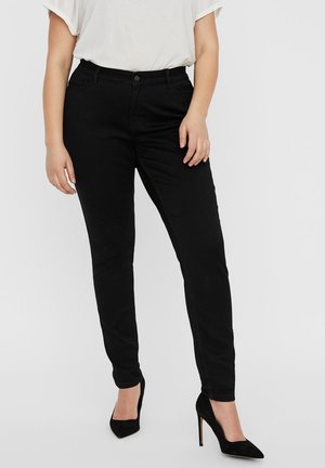 Person trägt enganliegende schwarze Hose, weißes kurzärmeliges Oberteil und schwarze High Heels, steht vor einem schlichten Hintergrund.