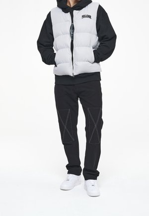 Gilet imbottito bianco sopra un hoodie nero, abbinato a pantaloni neri con accenti di cucitura a X. Sneakers bianche completano l'outfit.