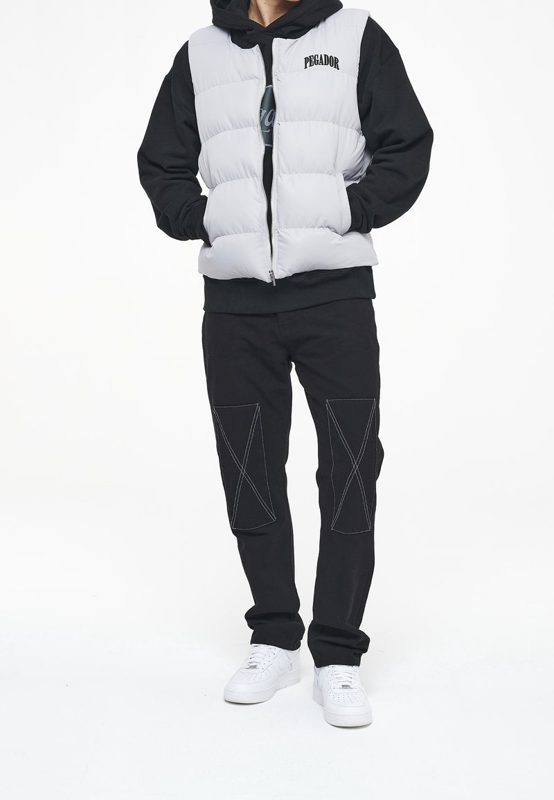 Vit vadderad väst över en svart hoodie, parat med svarta byxor med X-sömmar. Vita sneakers fulländar outfiten.