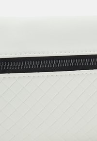 Biała torebka kopertowa z fakturą, z plecionym wzorem i czarnym suwakiem jako akcentem. Materiał wydaje się gładki i trwały.