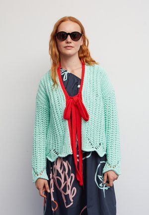 Minzgrüner Strickcardigan mit roter Krawatte, aus lockerem Strick mit gezacktem Saum, getragen über einem dunklen Kleid mit bunten abstrakten Mustern.