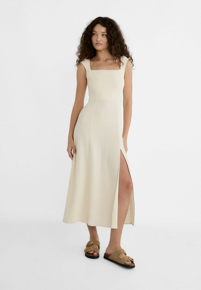 Beige linen midi dress with square neckline, ruffled shoulder details, and side slit. Model wears brown sandals with buckle accents.