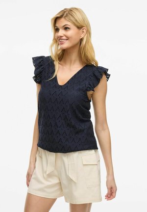 Haut bleu marine à col en V et manches volantes, présentant un motif à œillets. Associé à un short beige léger avec poches latérales.
