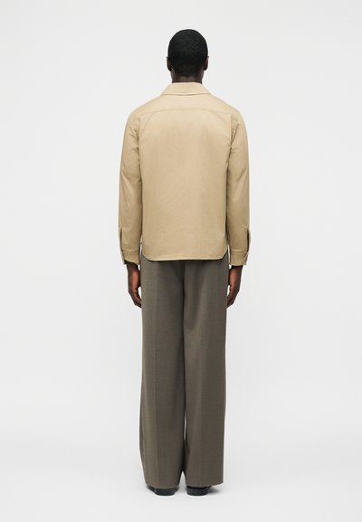 Άνδρας που στέκεται με την πλάτη γυρισμένη στην κάμερα, φορώντας μια μπεζ πουκάμισο με μακριά μανίκια και φαρδιά γκρι παντελόνια, σε ένα απλό φόντο.