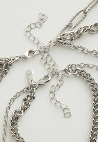 Srebrna metalna višeslojna ogrlica s lancima različitih veličina, višestrukim kopčama i teksturiranim naglascima između glatkih dijelova.