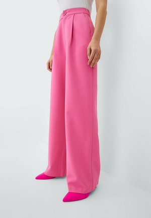 Frau trägt hoch taillierte, weit geschnittene rosa Hose mit Knopfdetail und spitze, knallpinke High Heels, stehend vor weißem Hintergrund.