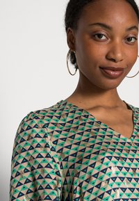 Green and beige blouse with geometric triangular pattern in dark blue and metallic accents; features a V-neck and puffed sleeves.