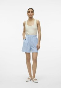 Canottiera leggera a coste beige abbinata a pantaloni corti blu e bianchi a righe, con vita elastica e tasche. Scarpe bianche senza lacci.